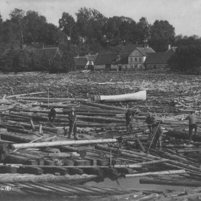 Skats pāri Mēmelei uz Sudmalu (tagad – Rīgas) ielas apbūvi no 54. līdz 60. namam. Priekšplānā sapostītā Plosta tilta atliekas, ko strādnieki mēģina atsvabināt no citiem kokmateriāliem. Tālāk starp stenceļiem atrodas kāda laiva ar nojumes konstrukciju.