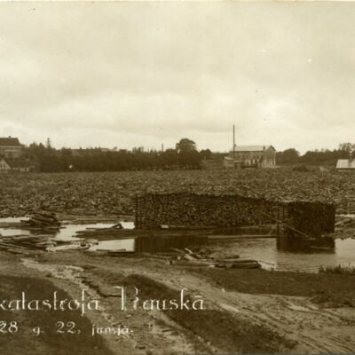 Skats pāri Mēmeles līkumam no tā saucamā “malkas plača” pie Sudmalu (tagad – Rīgas)  ielas 62. Priekšplānā sakrautas malkas grēdas, aiz tām tālumā redzamas Augšdzirnavas ar garu skursteni. Pa labi – Plosta kroga apbūve, pa kreisi – Sudmalu ielas ēkas.